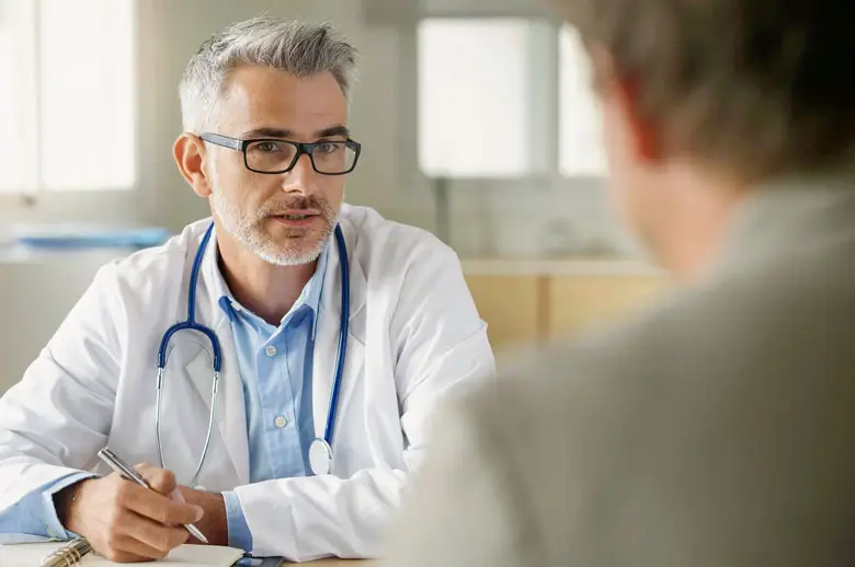 A middle-aged male physician with gray hair and glasses listens attentively while speaking with a patient in a clinical setting.