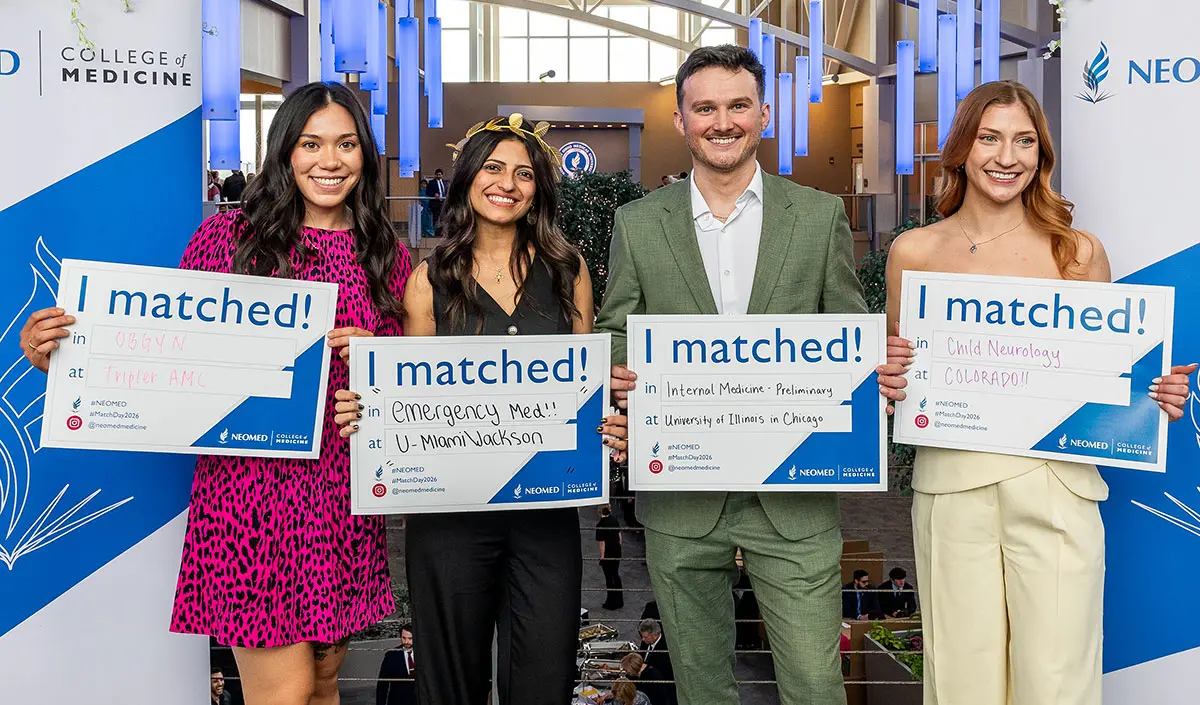 Four NEOMED students stand together holding “I matched!” signs with their residency placements, smiling in front of a NEOMED backdrop during a Match Day event.