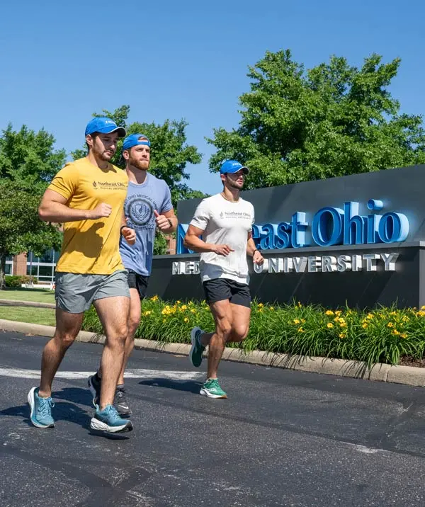 Three men in running gear run by the NEOMED sign on the NEOMED campus.