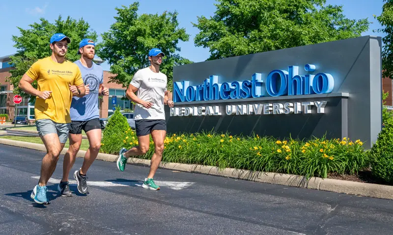 Three students jog past the NEOMED sign in front of the University.