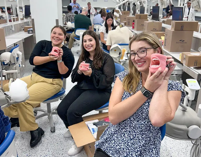 Three dental students in a simulation lab smile while holding model mouths with teeth, surrounded by dental equipment and boxes on their workstations.