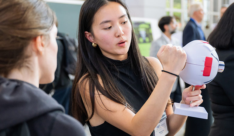 A woman holds a model eye while explaining her innovation at NEOvations Bed to Benchside event.