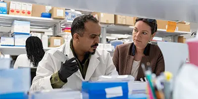 Two people in a laboratory discuss documents and equipment at a crowded workbench.