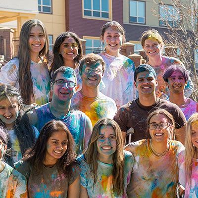 A joyful group of college students covered in vibrant color powder, celebrating together outdoors on a sunny day.
