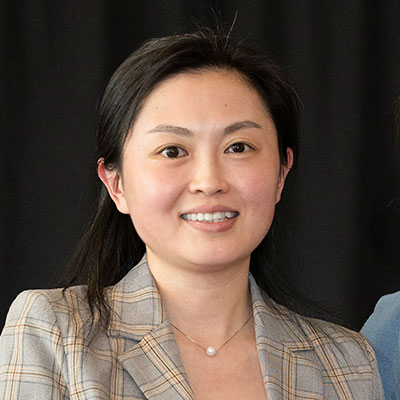 A confident woman in a plaid blazer smiles warmly against a neutral background, exuding professionalism and approachability.