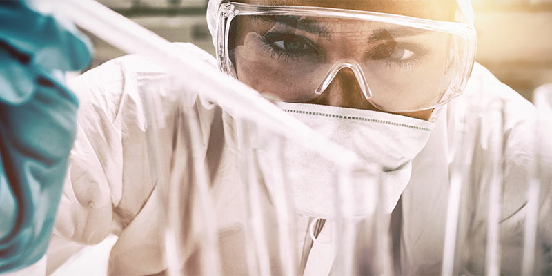 A researcher wearing safety goggles, gloves, and a face mask carefully examines test tubes in a laboratory setting.