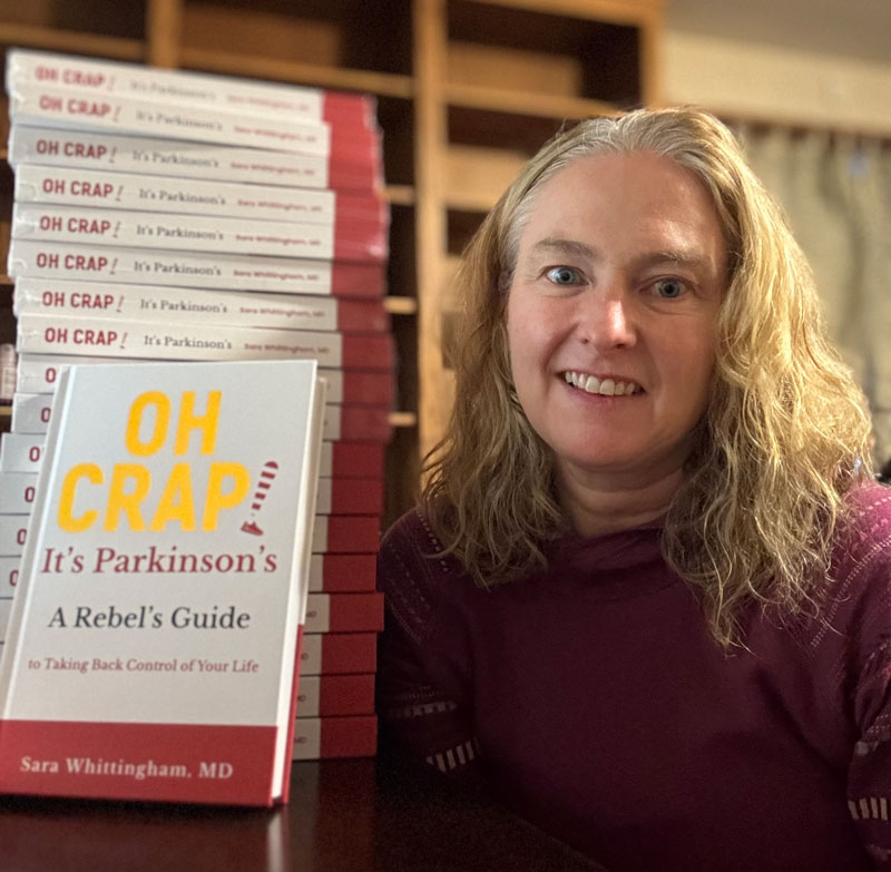 A woman sits beside a stack of books titled ‘OH CRAP! It’s Parkinson’s: A Rebel’s Guide to Taking Back Control of Your Life.’