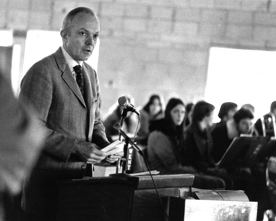 A man in a suit speaks at a podium before a seated audience.