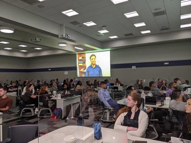 A group of people in a large room watch a presentation on screen.