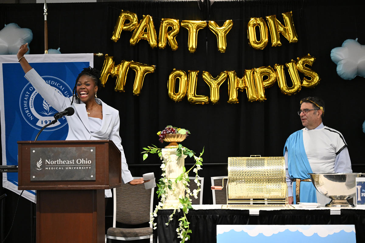 A speaker stands at a podium raising a hand during a Match Day event, with gold balloons reading “Party on Mt Olympus” and a raffle setup visible on stage.
