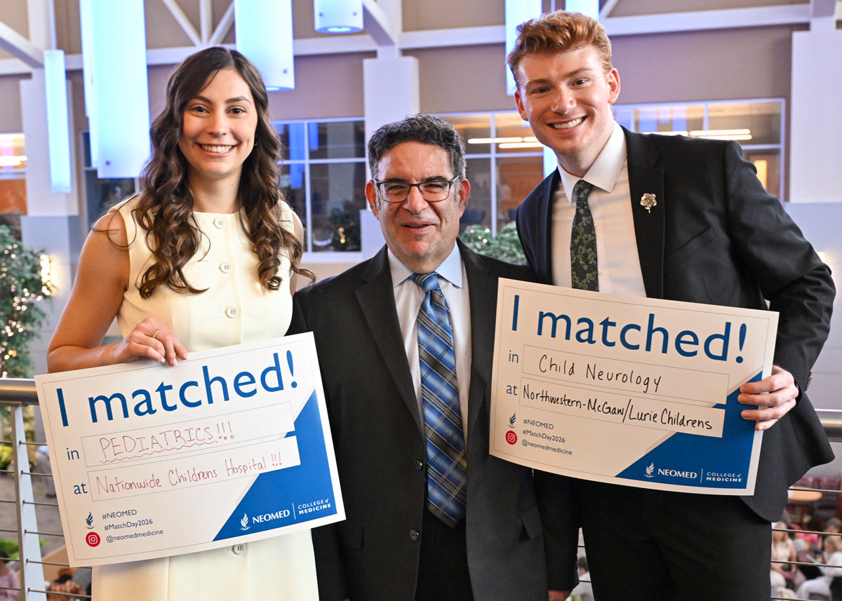 Three people pose together holding “I matched!” signs for pediatrics and child neurology placements, smiling in a bright atrium during a Match Day event.
