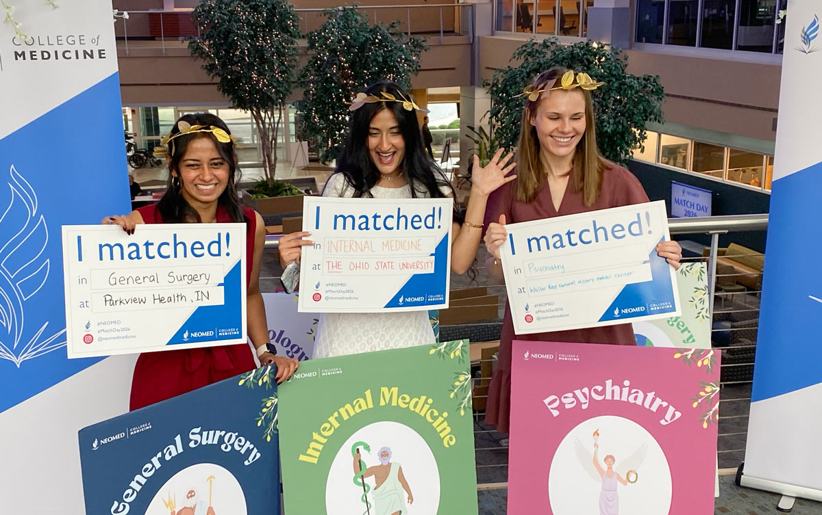 Three students hold “I matched!” signs showing their residency placements, smiling in a decorated indoor space during a Match Day event.