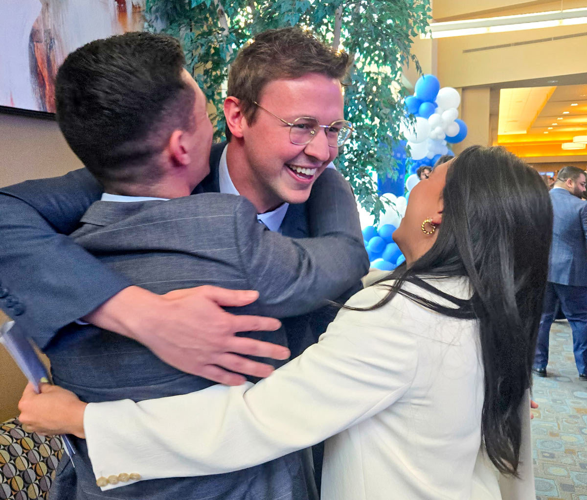 Three people embrace in celebration inside a large atrium, smiling and congratulating one another during a Match Day event.
