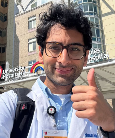 A student in his white coat gives a thumbs up while standing in front of the hospital where he is completing a rotation. 