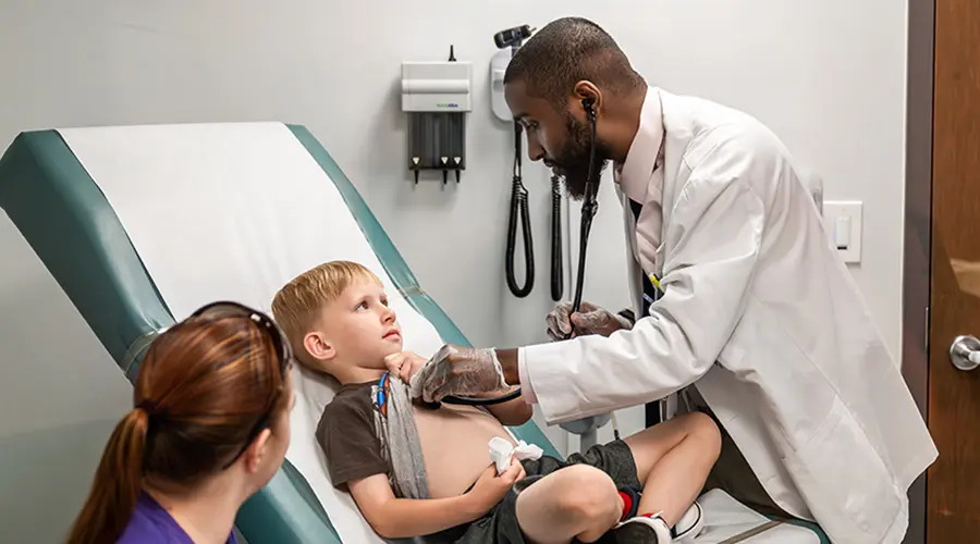 A student doctor examines a young boy with a stethoscope in a medical exam room, while the boy's mother holds his hand for comfort.