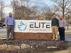 Three men stand beside a road sign that reads Elite Pharmacy.