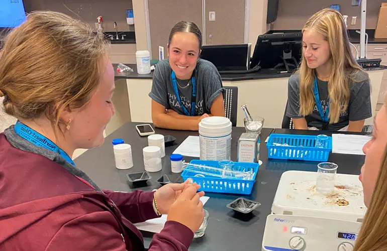 Four students sit around a lab table smiling and working together with test tubes, powders, and other science materials during a pharmacy-related activity.