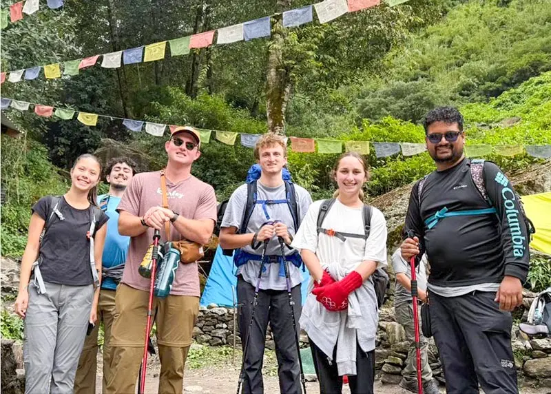 NEOMED students pose outdoors with hiking poles beneath colorful prayer flags, standing on a trail in a mountainous, forested landscape in Nepal.
