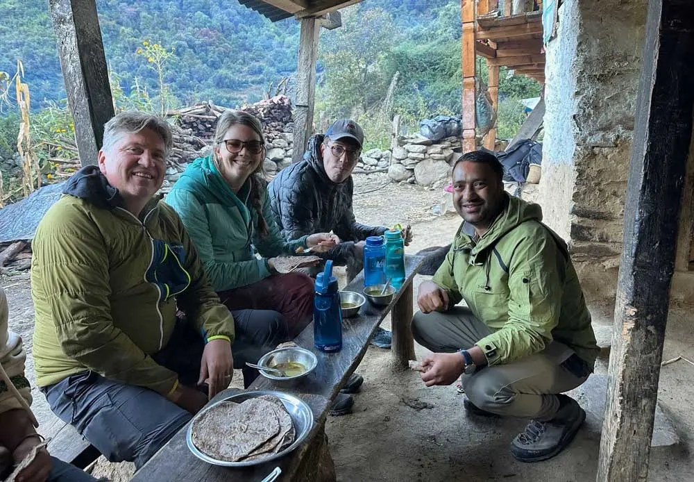 global-health-certificate-1000-01 A small group of adults sit on benches inside a rustic outdoor shelter, sharing a meal with metal plates and water bottles, with a mountainous landscape visible beyond the stone walls.