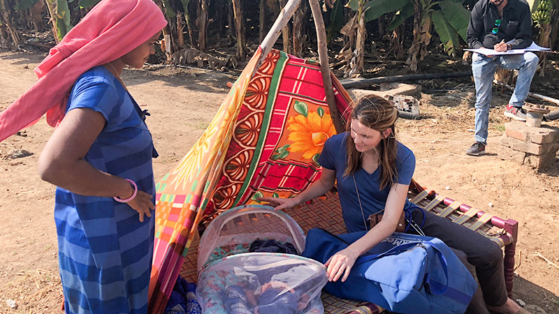 A student kneels beside a covered infant while speaking with a woman outdoors near a small shelter in a rural setting.