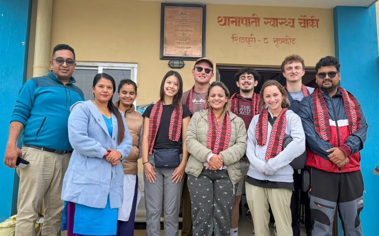 A group of students and community members stand together outside a health facility, wearing patterned scarves and smiling toward the camera.