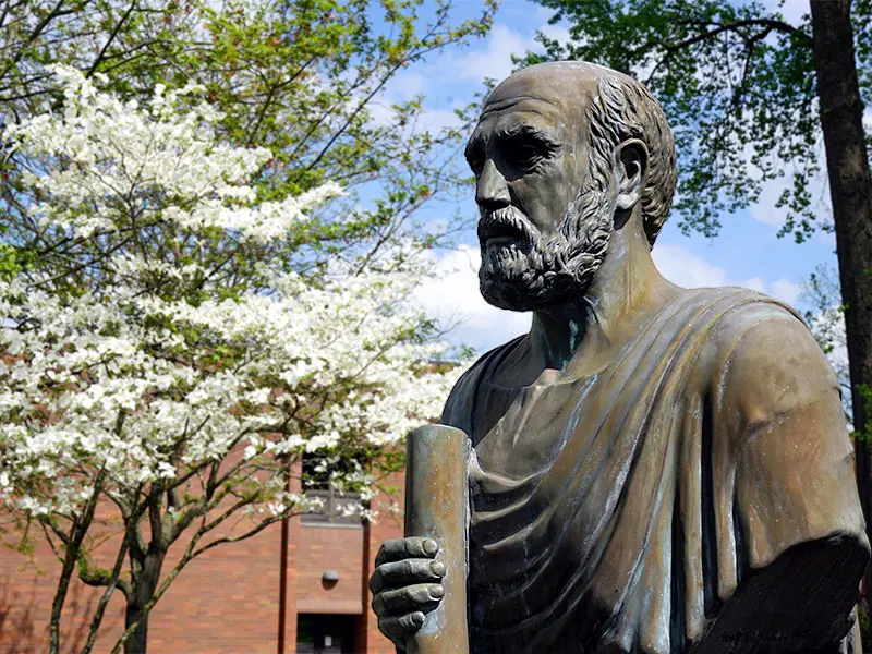 A bronze statue of Hippocrates on the NEOMED campus with flowering trees in the background.