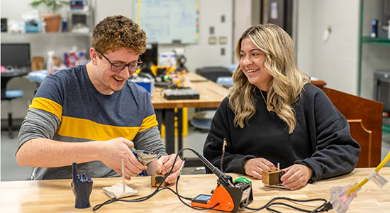 Two NEOMED students sit at a worktable smiling while assembling and testing electronic components in a laboratory setting.