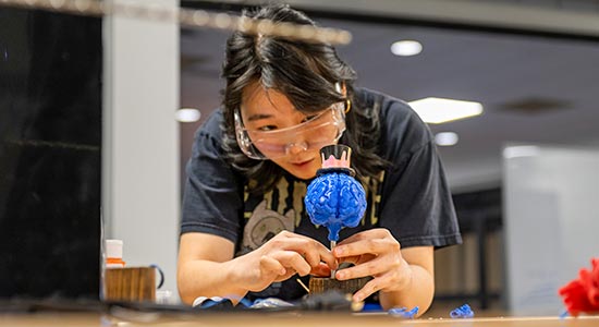 A student wearing safety glasses carefully adjusts a small blue prototype device on a worktable indoors.