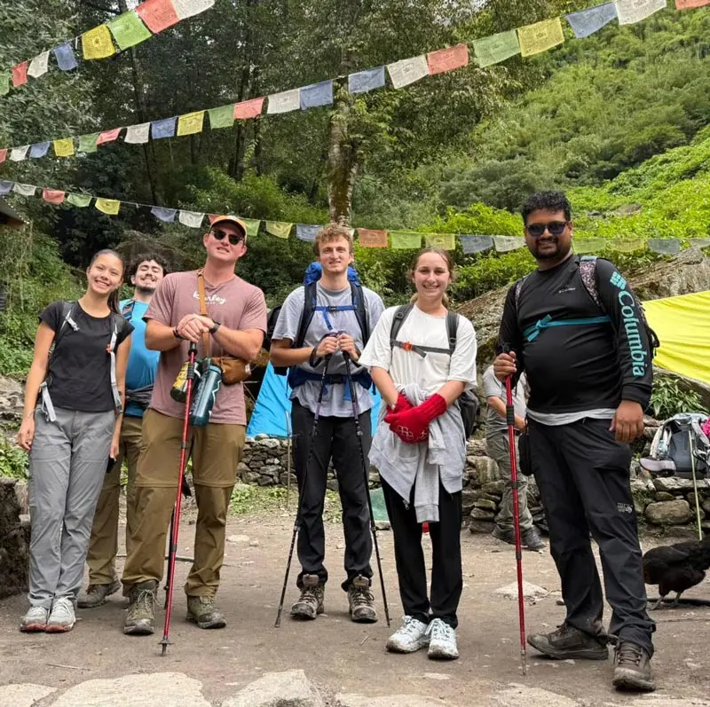 NEOMED students pose outdoors with hiking poles beneath colorful prayer flags, standing on a trail in a mountainous, forested landscape in Nepal.