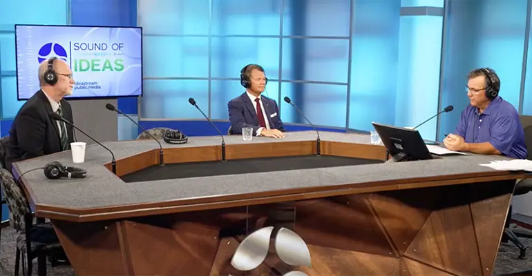 Three men sit at a broadcast studio table wearing headsets and speaking into microphones during a live recording of the radio program Sound of Ideas.
