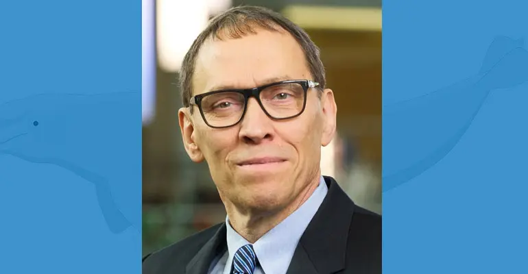 A person wearing a suit and glasses stands against a blue background that includes a diagram of a whale.