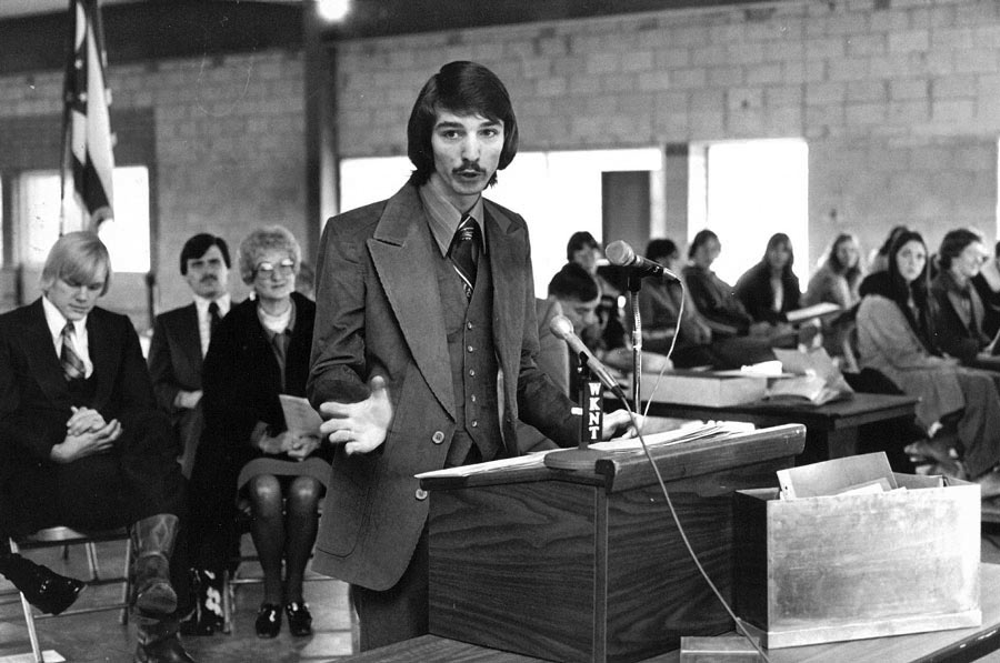 A young man in a suit speaks at a podium before a seated audience.